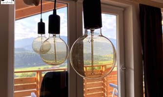 Three hanging light bulbs in front of a large window overlooking a green mountain landscape. The view shows a bright sky and a terrace in the background.