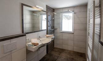 A modern bathroom with a sink, mirror, and a shower. Bright tiles and daylight create a friendly atmosphere.
