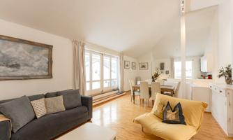 A modern living room with bright furnishings and wooden flooring. There are large windows that let in plenty of light, as well as a dining area and a cozy seating corner.