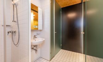 A modern bathroom with a shower, a sink, and a large mirror. The walls are mostly tiled in white, with a paneled wooden ceiling and black partitions.