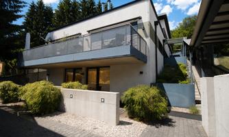 A modern house with a balcony and large window front. The surroundings are green and well-maintained, ideal for relaxing moments.