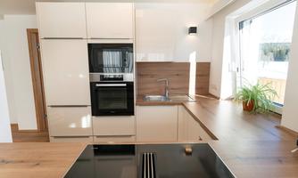 A modern kitchen with a bright design and wood accents. It features an oven, a cooktop, and a window with a plant.