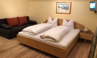 A cozy room with a double bed and two pillows. On the left side, there is a couch with cushions and a TV on the wall.