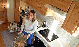 A woman takes a tray of freshly baked food out of the oven in a bright kitchen. The woodwork and modern furnishings create an inviting atmosphere.