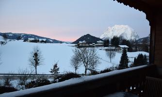 A winter landscape with snow-covered fields and mountains in the background. The sky displays soft colors in the twilight.