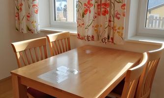 A bright dining area with a wooden table and four chairs. The windows are adorned with colorful curtains.