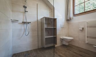 A modern bathroom with a shower, a toilet, and a wooden shelf. Large windows provide plenty of daylight.