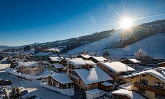 A winter landscape with snow-covered houses and mountains in the background. The sun rises on the horizon and the sky is clear and blue.