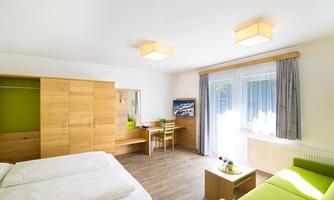 A modern, bright room with two beds, a table, and a sofa. The furniture is made of wood with green accents and large windows.