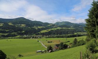 A picturesque landscape with green meadows and gentle hills. In the background, mountains and a clear sky can be seen.
