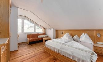 A cozy bedroom with a double bed and a seating area. The wooden paneling and large windows create a bright, inviting atmosphere.