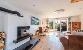 A modern living room with a fireplace, a television, and large windows. There is a dining table and a cozy seating area with a comfortable armchair.