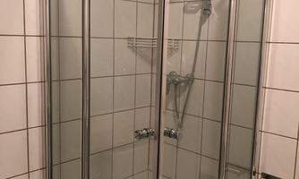 A modern shower cabin with glass walls and white tiles. The shower has a hand shower and a practical shelf.