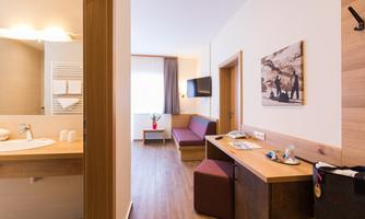 A modern hotel room with a cozy living area and a bathroom. The bright decor and wooden details create an inviting atmosphere.