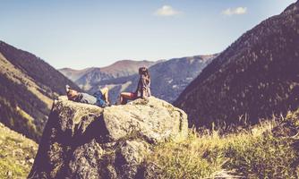 Two people are relaxing on a large rock in the mountains. In the background, gentle hills and a clear sky are visible.