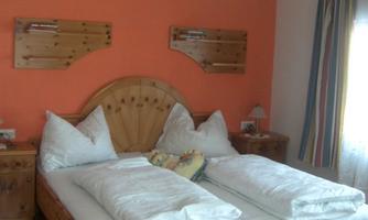 A cozy bedroom with a wooden double bed and white bedding. The walls are orange and there are some wooden furniture as well as a colorful curtain at the window.
