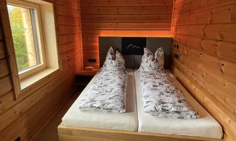 A cozy wooden bedroom with two single beds and modern pillows. The warm light and the large window create a relaxed atmosphere.