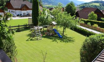 A beautiful garden with lawn, trees, and a playground. In the background, rooftops and mountains are visible.