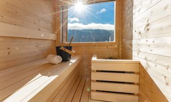 A bright sauna with wooden paneling and a window through which the sun shines. On the bench lies a towel and in the corner stands a bucket with a wooden ladle.