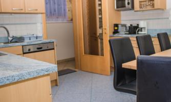 A modern kitchen with wooden cabinets and a dining table. In the background, an open door and some kitchen appliances are visible.