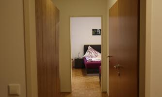 A hallway with two closed doors and an open door that leads to a bedroom. In the bedroom, there is a bed with colorful pillows and a nightstand.