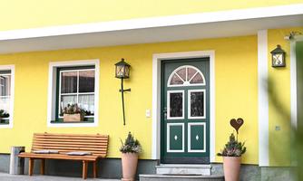 A yellow hunting lodge with a green door and a landscaped entrance area. In front of the house is a bench and there are decorative elements such as plants and a trophy.