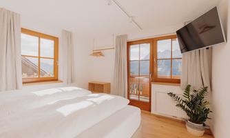 A cozy bedroom with two beds, large windows, and a television. The decor is modern and bright, with a beautiful view of the mountains.