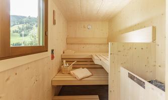 A bright sauna room with wooden shelves and a window to the outside area. On the lounger, there are towels and a basket with sauna accessories.