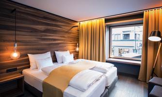 A modern hotel room with a double bed and soft pillows. Large windows let in plenty of light, and the warm colors create an inviting atmosphere.