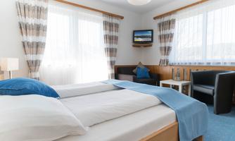 A cozy room with a double bed, a seating area, and a small television. The room has large windows and bright curtains that let in a lot of daylight.