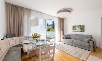 A modern living room with a cozy gray sofa and a wooden dining area. Large windows let in plenty of daylight and a bouquet of flowers adds color.