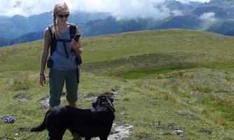 A woman is standing with her dog on a green meadow in the mountains. In the background, cloud-covered mountains are visible.
