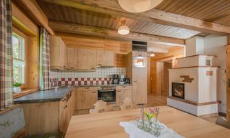 A cozy wooden kitchen with a dining table and a fireplace. The windows are decorated with checkered curtains, and fresh flowers adorn the table.