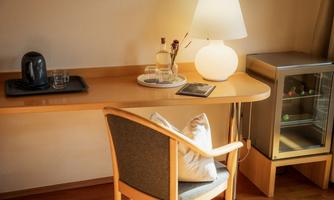 A cozy desk with a chair, a lamp, and a small fridge. Drinks and a tablet are on the table.