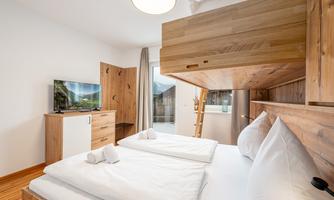 A modern bedroom with two beds and a wooden loft bed. There is a television and a bright, inviting atmosphere.