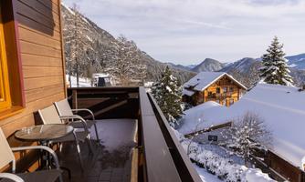 A balcony with two chairs and a table, surrounded by snow-covered mountains and wooden houses. The sky is clear and the landscape is wintery beautiful.