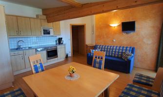 A modern kitchenette with wooden shelves and a dining area. In the background, there is a blue sofa bed and a television is mounted on the wall.