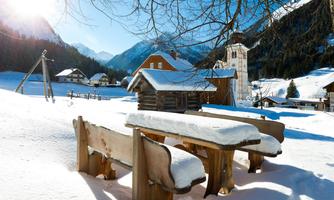 A winter landscape with a wooden table and benches covered in snow. In the background, you can see mountains and a picturesque village with a church.