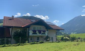A charming house with a red roof and flower balcony in a green meadow landscape. In the background, impressive mountains and a clear blue sky can be seen.