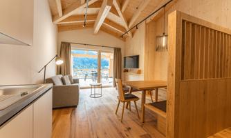 A modern living room with wooden floorboards and a large glass front. The room is bright and friendly, featuring a kitchen, a dining table, and cozy seating.