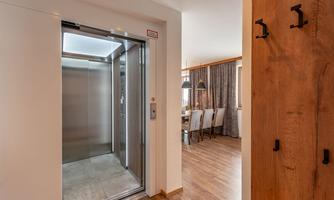 A modern elevator in a bright hallway. In the background, there is a dining area with a table and chairs, as well as windows with curtains.