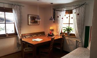 A cozy dining area with a wooden table and chairs. Foreground: a plant and a window with curtains, through which daylight falls.