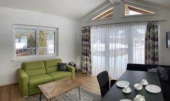 A modern living room with a green couch and a dining area. Large windows provide a view of the snowy landscape outside.