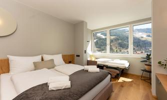 A modern bedroom with a double bed and a large window that offers a view of the mountains. The decor is bright and friendly, with a cozy space for relaxation.