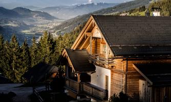 A cozy wooden house in a mountainous landscape. In the background, the Alps and a wooded valley can be seen.