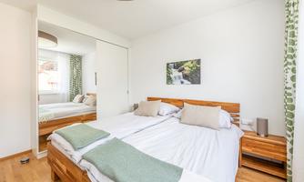 A bright bedroom with two single beds and wooden furniture. In the background, there are curtains with a green pattern and a picture of a waterfall.
