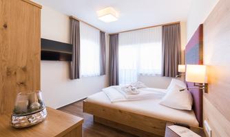 A modern bedroom with wooden furniture and a large bed. Light floods the room through the windows with curtains.