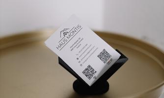 A black card holder for a business card stands on a golden table. The business card displays the name "HAUS MONTIS" and contains a QR code.