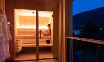 A modern sauna with warm wooden interiors and soft lighting. One person sits relaxed inside while it is dark outside.