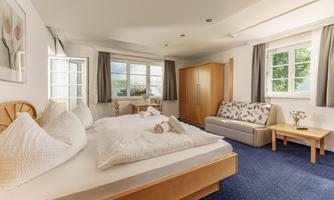 A bright room with two beds, a sofa, and a table. Large windows let in a lot of light.